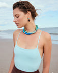 Woman wearing a light blue top and brown pants with a blue necklace on a beach.
