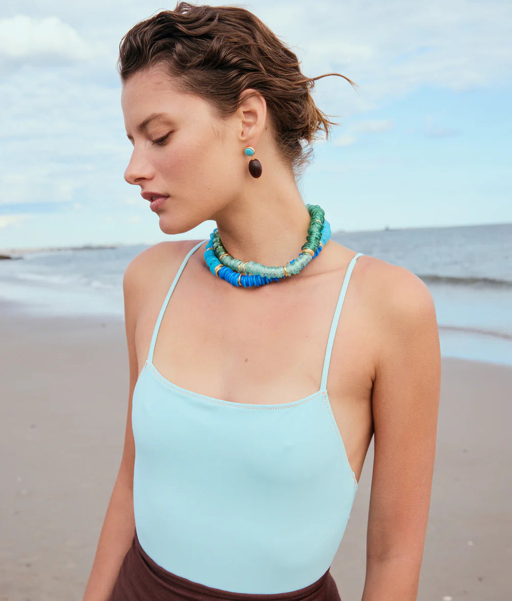 Woman wearing a light blue top and brown pants with a blue necklace on a beach.
