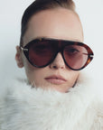 model wearing tortoise sunglasses with gold accents and light red lenses on a grey background 