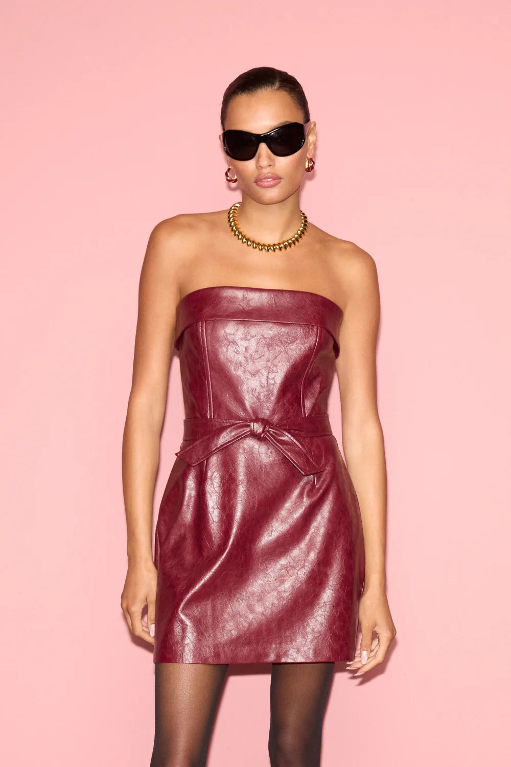 A model wearing a strapless burgundy faux-leather mini dress with a matching tie belt, styled with black sunglasses, a gold chain necklace, and black tights against a solid pink background.