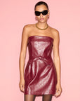 A model wearing a strapless burgundy faux-leather mini dress with a matching tie belt, styled with black sunglasses, a gold chain necklace, and black tights against a solid pink background.