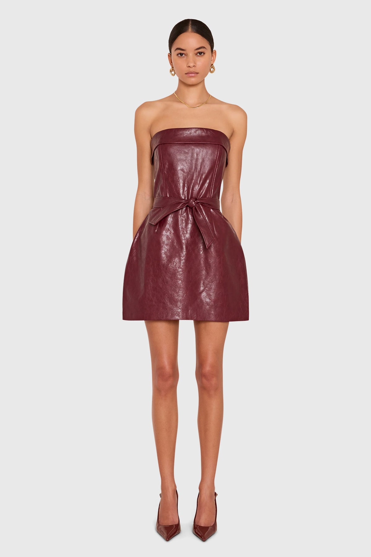 A model wearing a strapless, shiny burgundy faux-leather mini dress with a tie waist, paired with matching pointed-toe heels, against a light gray background.