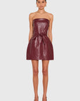 A model wearing a strapless, shiny burgundy faux-leather mini dress with a tie waist, paired with matching pointed-toe heels, against a light gray background.