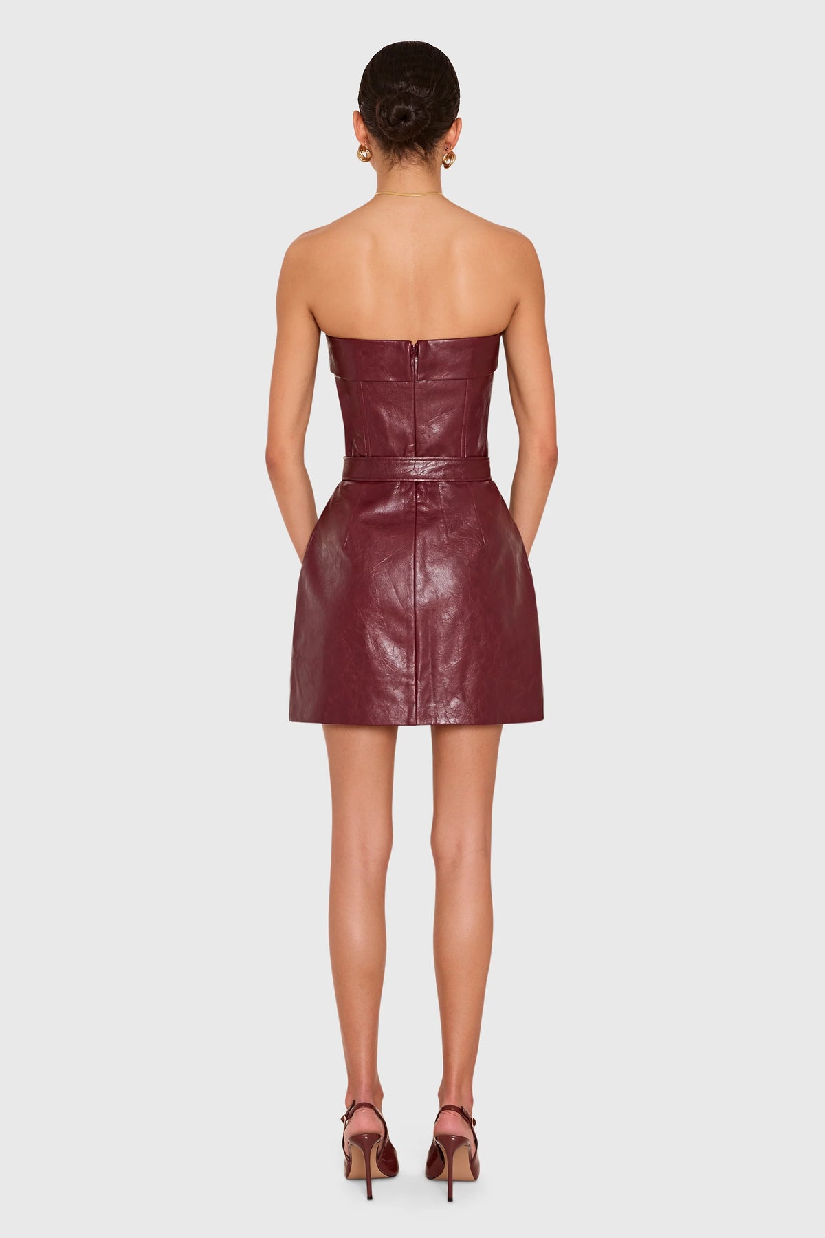 A woman viewed from the back wearing a strapless, fitted mini dress in a deep burgundy faux leather, paired with matching burgundy high-heeled sandals, against a light gray studio background.