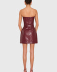 A woman viewed from the back wearing a strapless, fitted mini dress in a deep burgundy faux leather, paired with matching burgundy high-heeled sandals, against a light gray studio background.