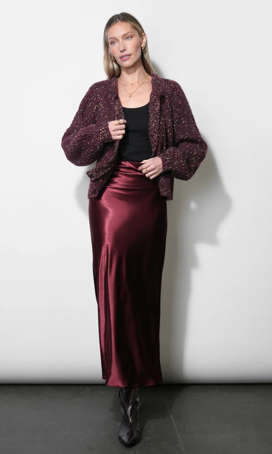 A woman modeling a dark burgundy textured knit cardigan over a black top and a matching satin midi skirt, paired with black ankle boots, against a plain light gray studio background.