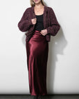 A woman modeling a dark burgundy textured knit cardigan over a black top and a matching satin midi skirt, paired with black ankle boots, against a plain light gray studio background.