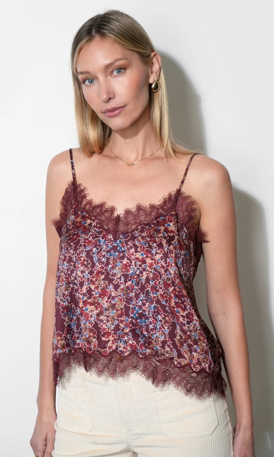 A woman wearing a burgundy lace camisole top with a red, blue, and white floral print, paired with cream-colored pants, standing against a plain white wall.