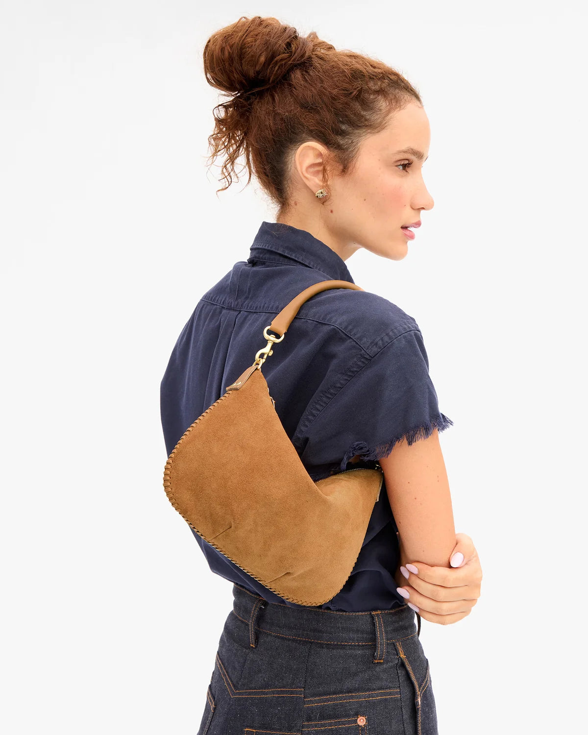 A woman shown from the back wearing a brown suede crescent shoulder bag with gold-tone hardware, styled with a navy shirt and dark denim jeans against a plain white background.