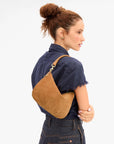 A woman shown from the back wearing a brown suede crescent shoulder bag with gold-tone hardware, styled with a navy shirt and dark denim jeans against a plain white background.