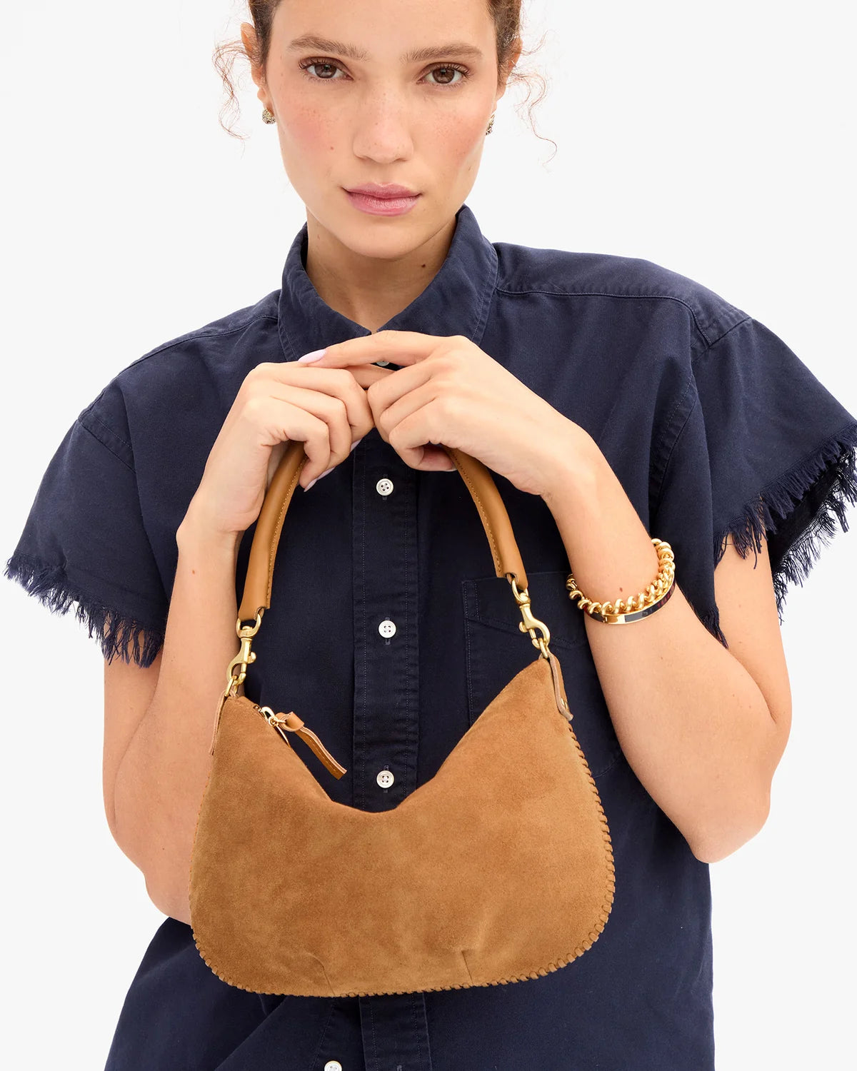 A woman wearing a navy blue short-sleeve button-up shirt holds a tan suede crescent shoulder bag with gold-tone hardware against a plain white background.