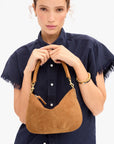 A woman wearing a navy blue short-sleeve button-up shirt holds a tan suede crescent shoulder bag with gold-tone hardware against a plain white background.