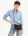 A model wearing a light blue denim shirt and jeans, carrying a dark brown suede crescent-shaped crossbody bag with a black strap and gold-tone hardware against a plain white background.