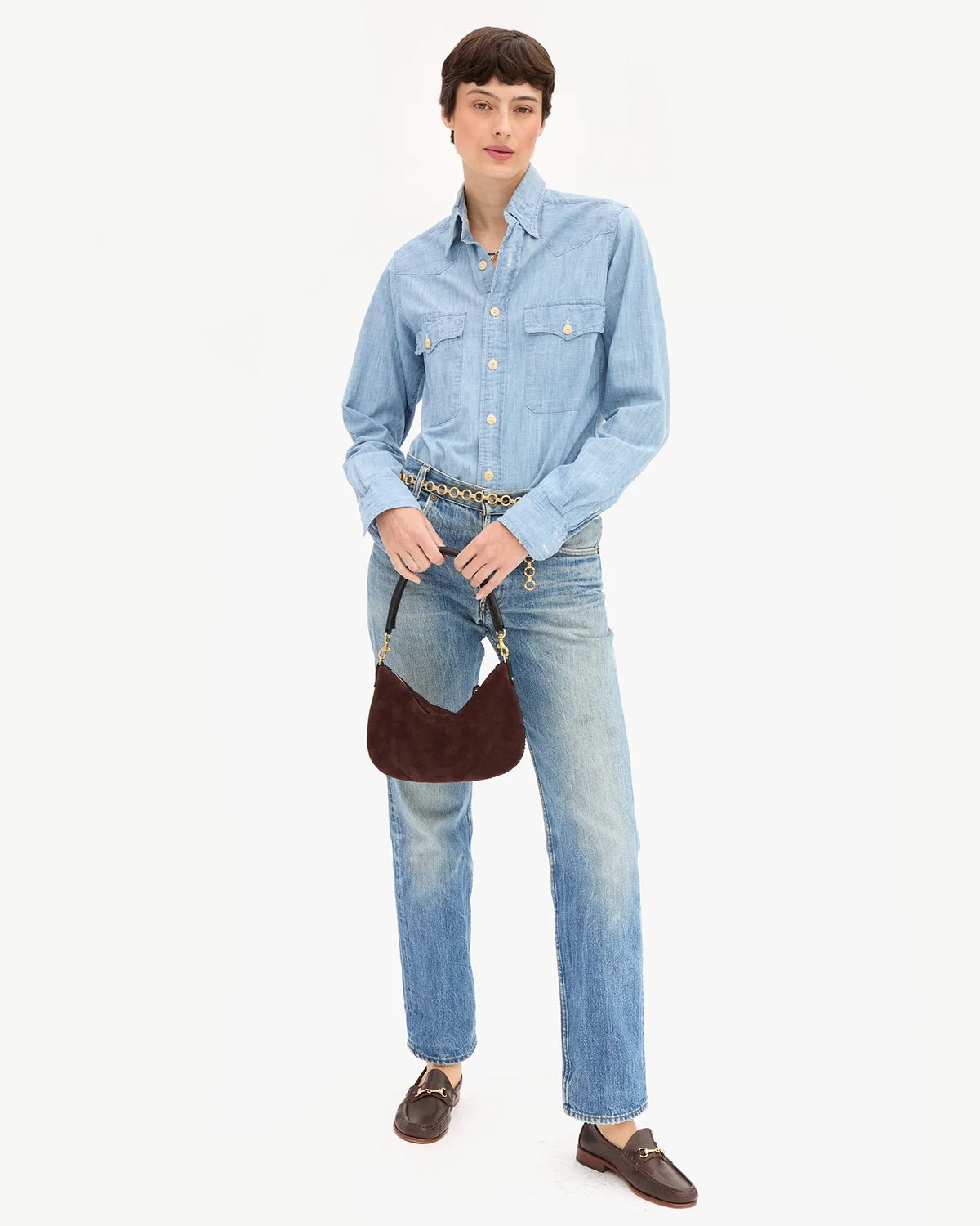 A model stands against a plain white studio background wearing a light blue denim button-up shirt, light blue denim jeans, a patterned belt, dark brown leather loafers, and holding a dark brown crescent shoulder bag.