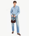 A model stands against a plain white studio background wearing a light blue denim button-up shirt, light blue denim jeans, a patterned belt, dark brown leather loafers, and holding a dark brown crescent shoulder bag.