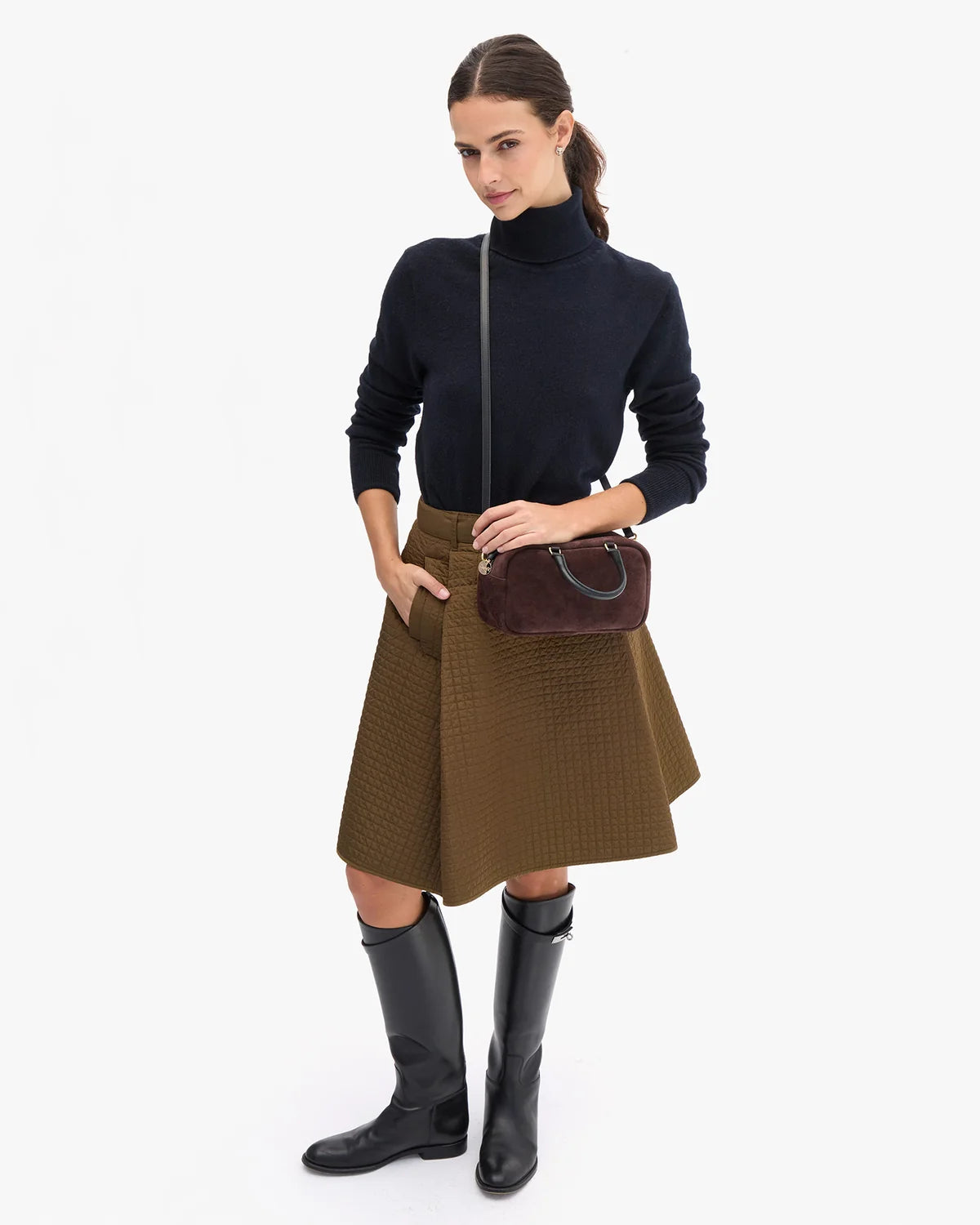 A woman modeling a black turtleneck sweater, a brown textured pleated skirt, black knee-high boots, and a dark brown crossbody handbag, photographed against a clean white studio background.