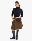 A woman modeling a black turtleneck sweater, a brown textured pleated skirt, black knee-high boots, and a dark brown crossbody handbag, photographed against a clean white studio background.