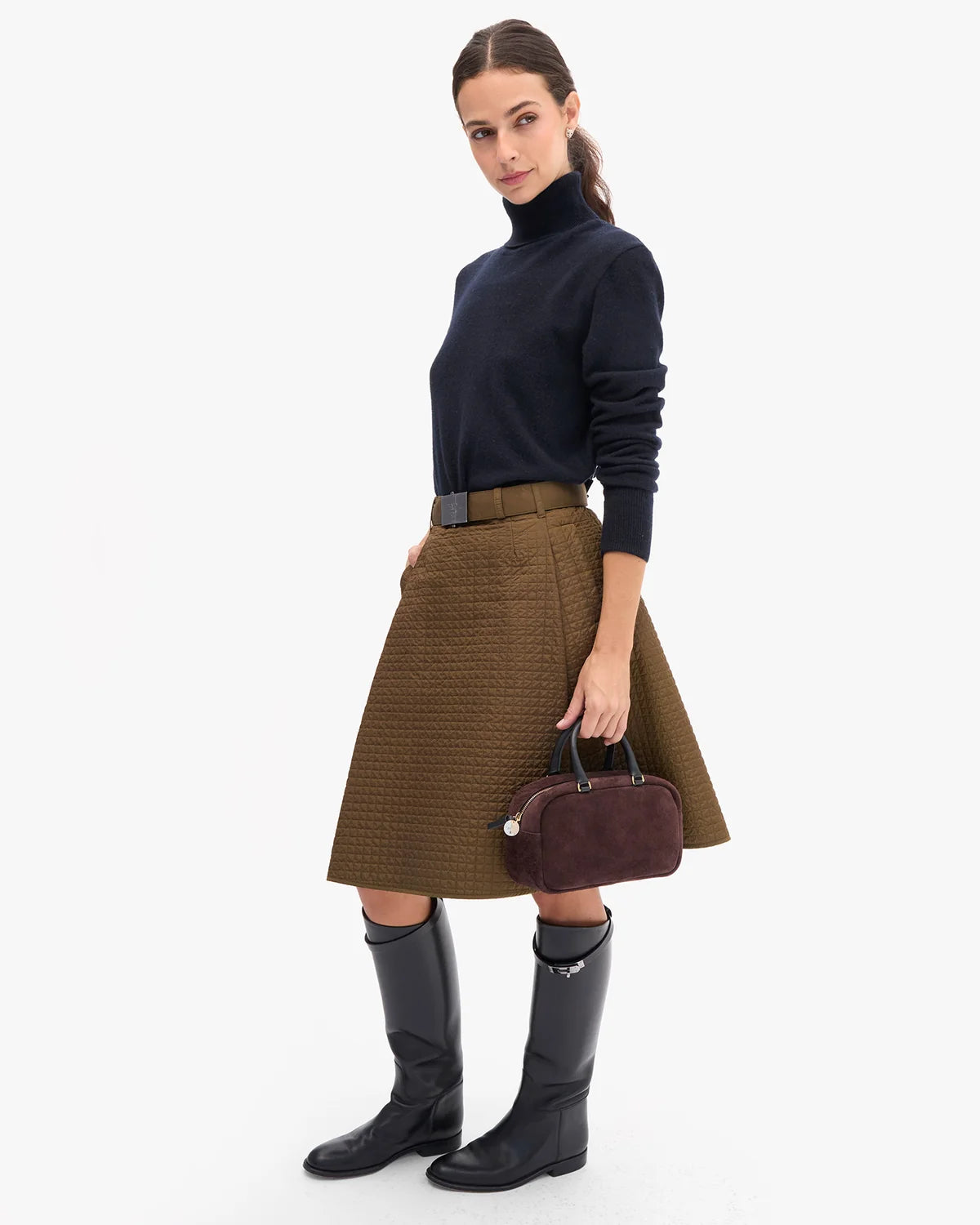 A woman models a brown textured pleated skirt with a black belt, paired with a navy turtleneck, black knee-high boots, and a dark plum handbag, set against a plain white studio background.