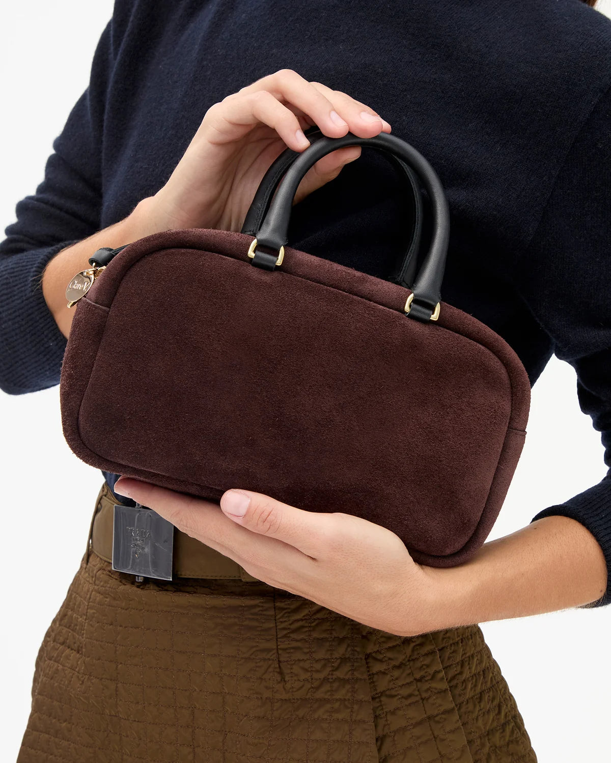 A person holding a small structured handbag in dark brown suede with black leather handles and gold-tone hardware, shown against a clean white background.