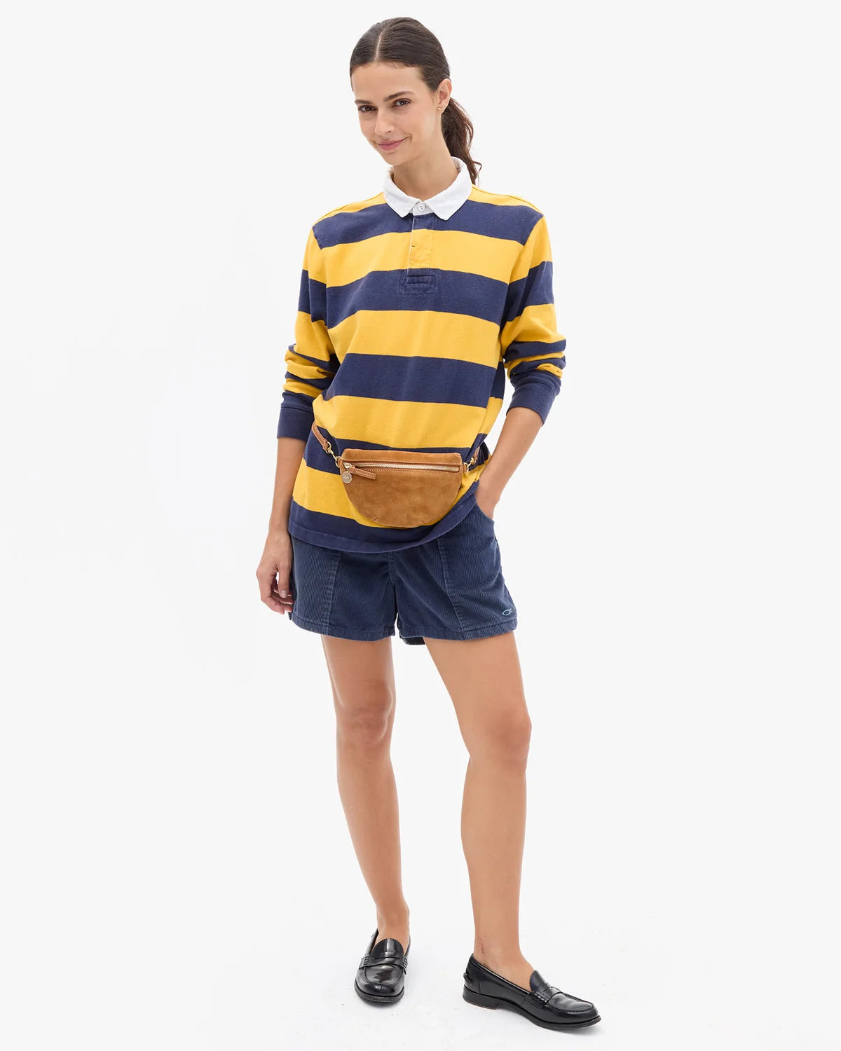 A woman modeling a yellow and navy blue horizontally striped long-sleeve rugby shirt with a white collar, paired with navy shorts, a brown belt bag, and black loafers, against a plain white background.