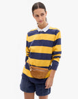 A woman modeling a yellow and navy blue horizontally striped long-sleeve rugby shirt with a white collar, paired with navy shorts and a brown belt bag, against a light gray background.