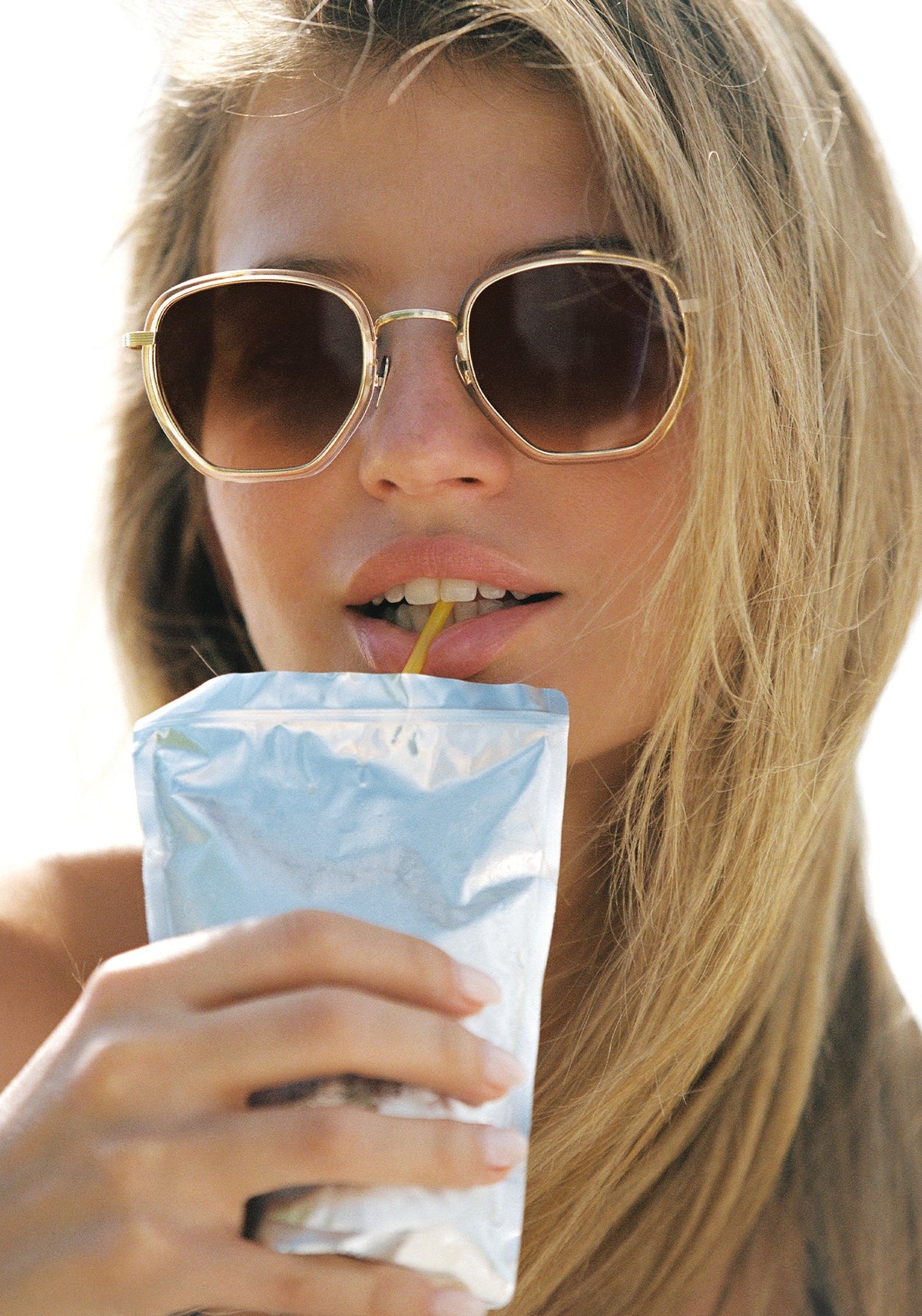 A close-up lifestyle shot of a woman with blonde hair wearing gold-framed sunglasses and drinking from a light blue foil pouch with a yellow straw against a bright white background.