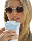 A close-up lifestyle shot of a woman with blonde hair wearing gold-framed sunglasses and drinking from a light blue foil pouch with a yellow straw against a bright white background.