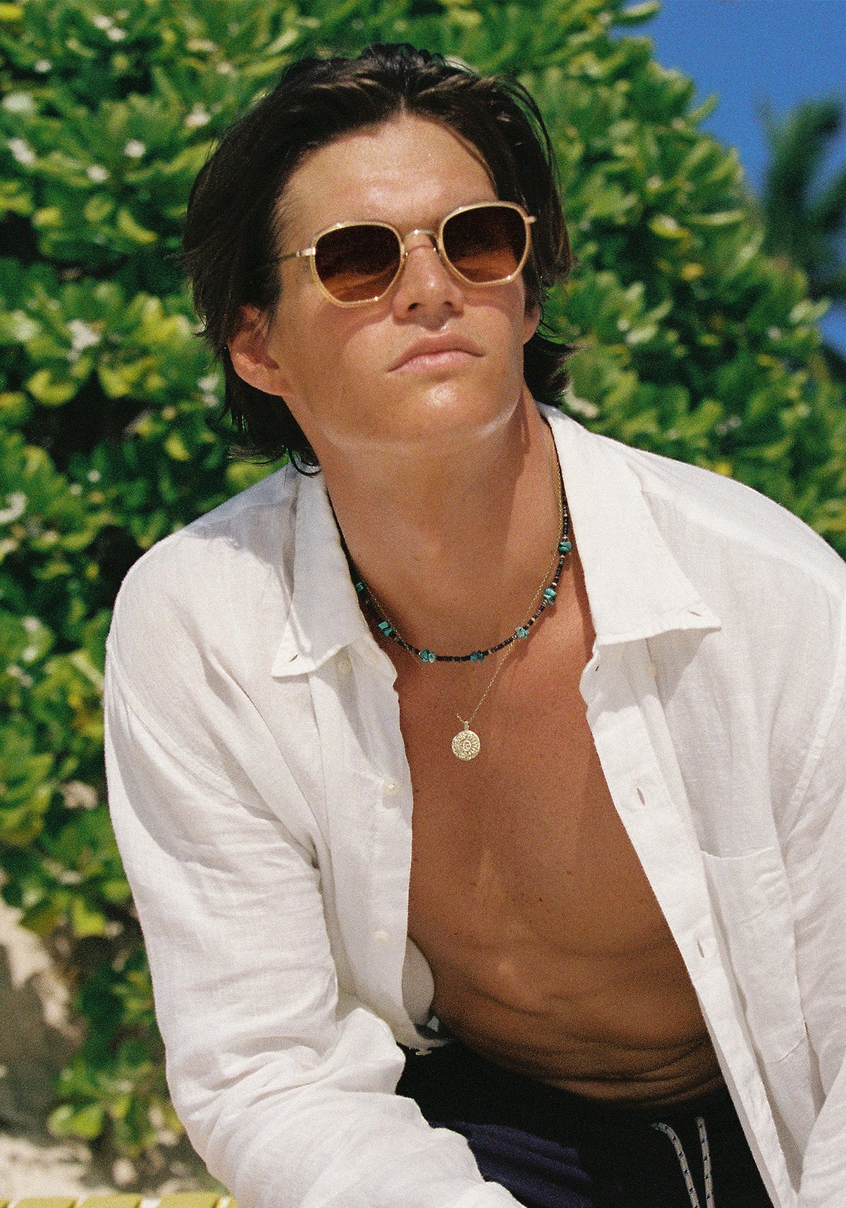 A man wearing gold-framed sunglasses, a white button-down shirt, and layered necklaces, posed outdoors in front of green foliage.