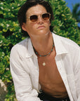 A man wearing gold-framed sunglasses, a white button-down shirt, and layered necklaces, posed outdoors in front of green foliage.