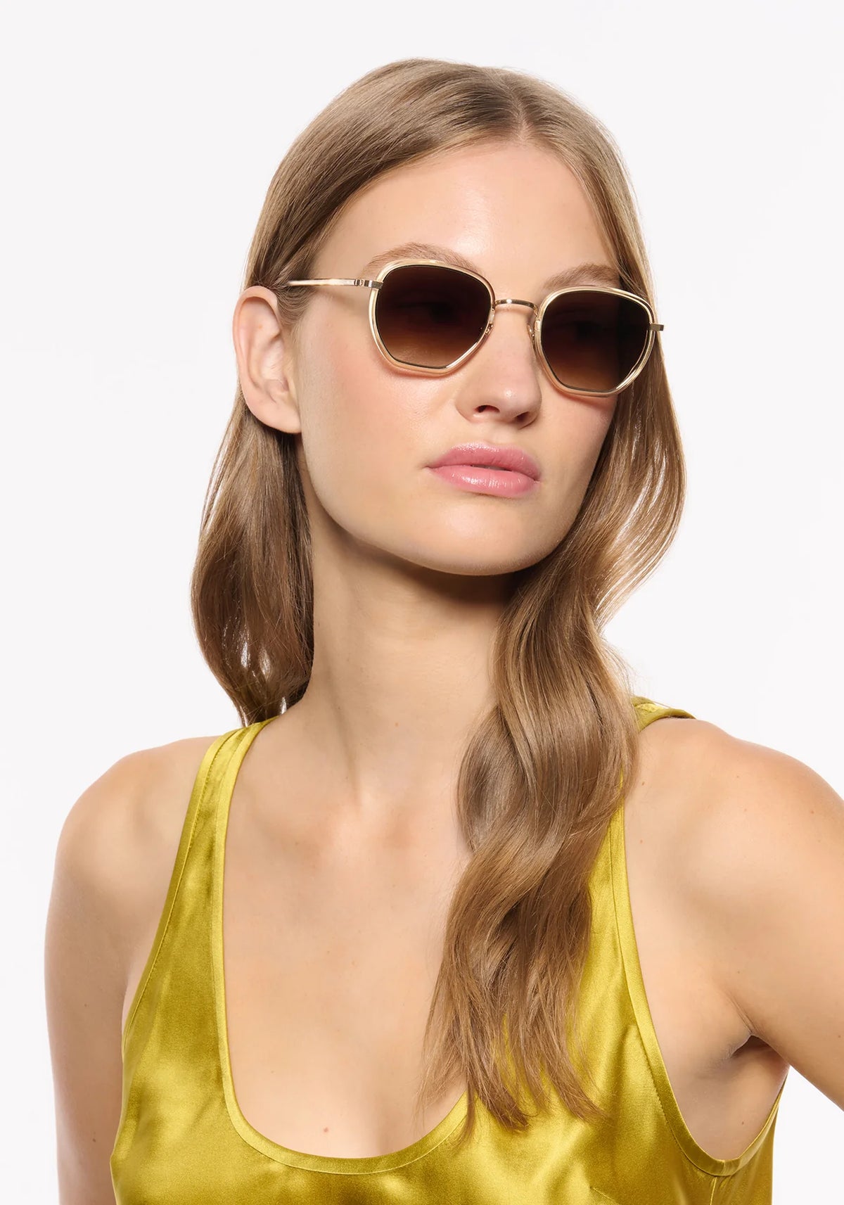 A woman with light brown hair wearing round gold-framed sunglasses with dark brown lenses and a shiny gold satin top, photographed against a plain white background.