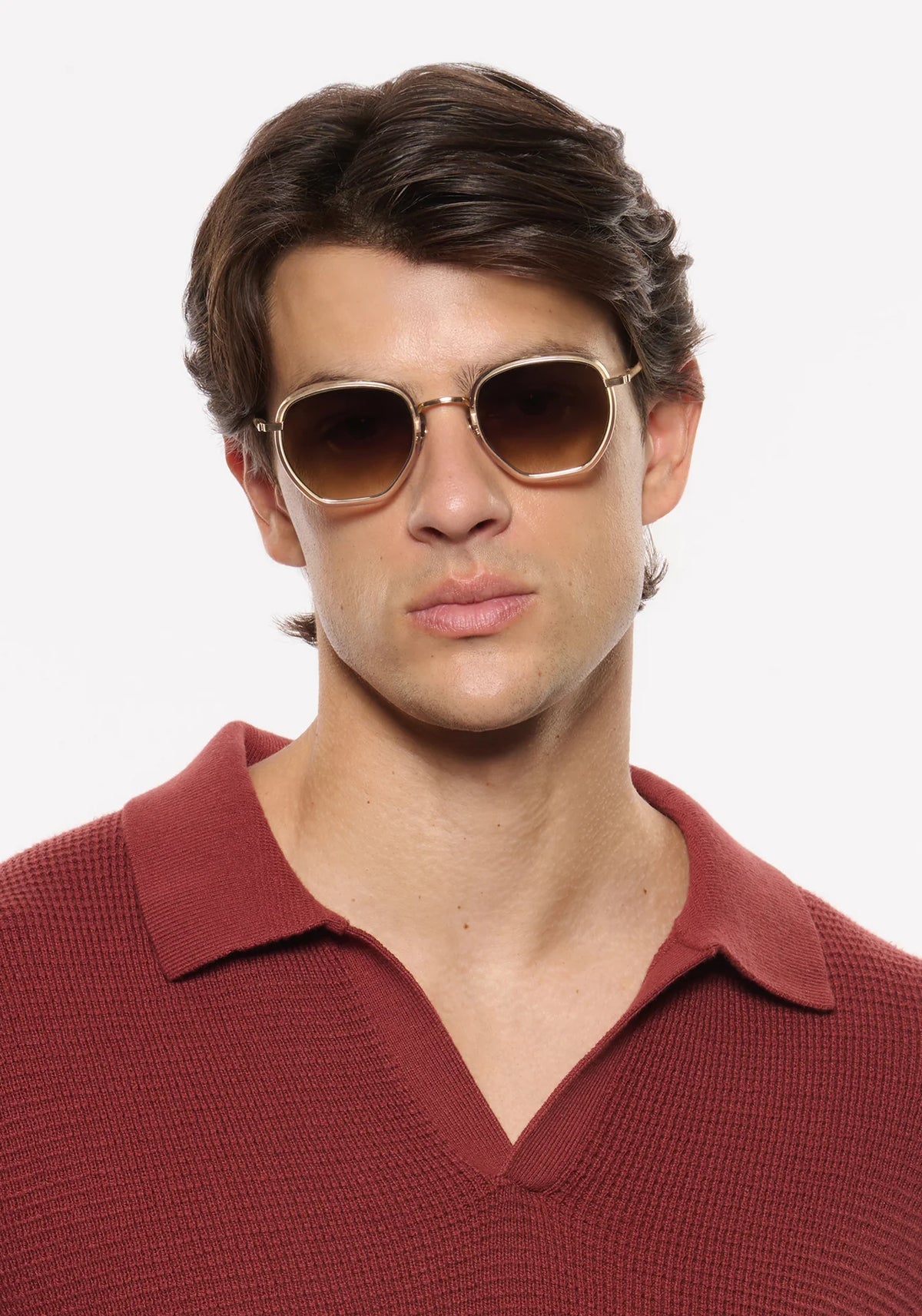 A man wearing round gold-framed sunglasses with brown lenses and a textured burgundy polo shirt, photographed against a clean white background.