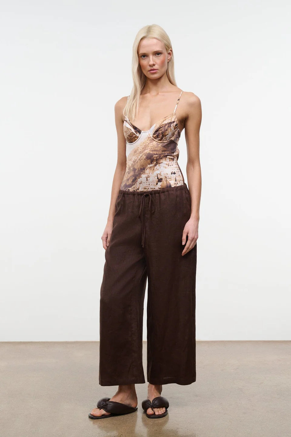 A blonde model stands against a plain white studio wall wearing a sleeveless camisole top with a brown and beige abstract print, paired with dark brown wide-leg pants and black slide sandals.