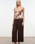 A blonde model stands against a plain white studio wall wearing a sleeveless camisole top with a brown and beige abstract print, paired with dark brown wide-leg pants and black slide sandals.