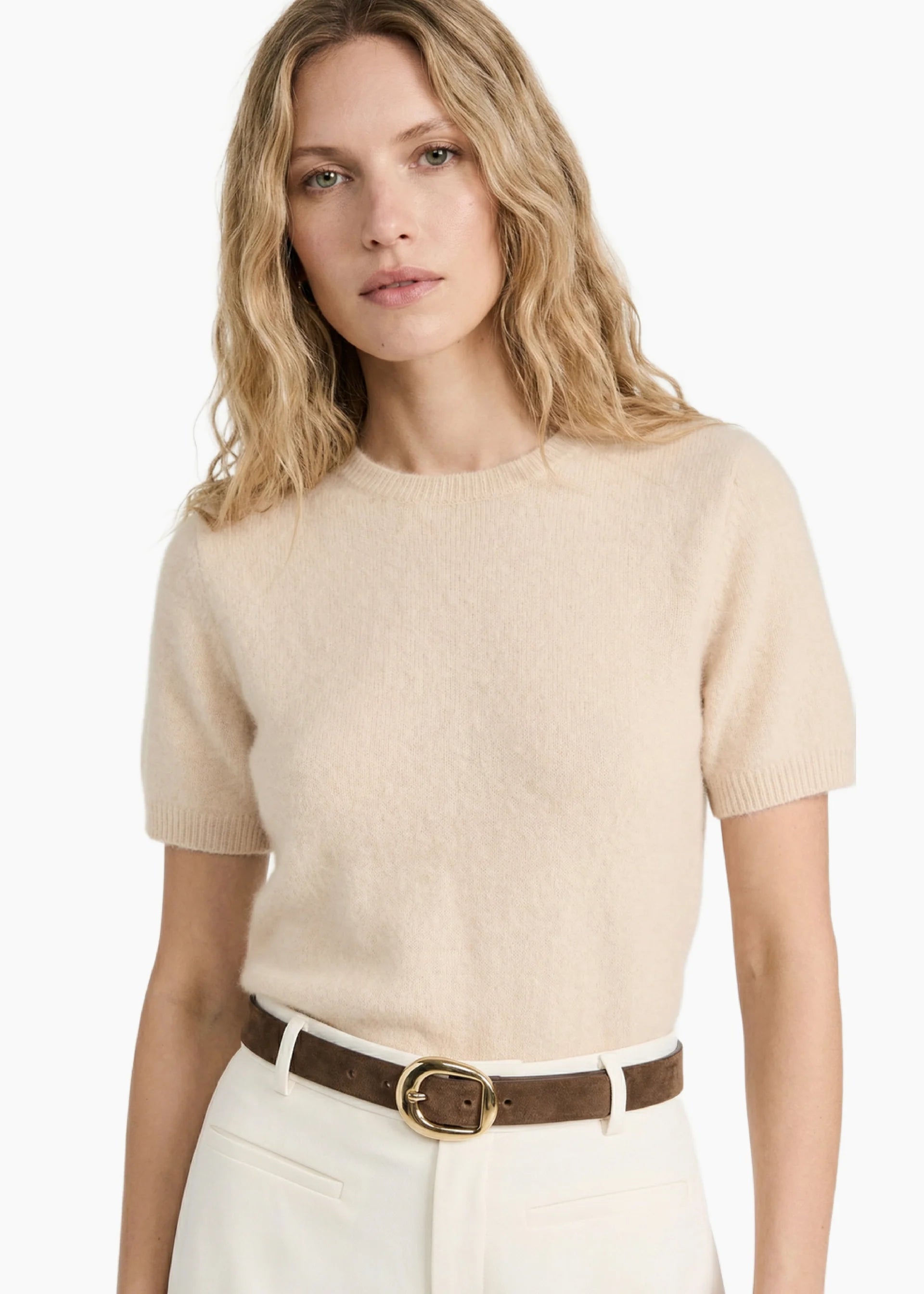Woman wearing a beige sweater and white pants with a brown belt on a white background