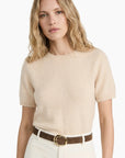 Woman wearing a beige sweater and white pants with a brown belt on a white background