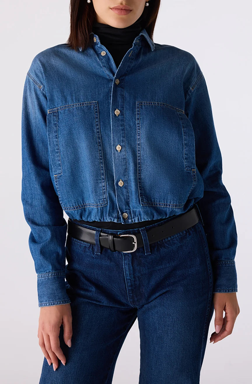 A model wearing a cropped, long-sleeve blue denim shirt with buttoned chest pockets, tucked into dark blue jeans with a black belt, against a plain light gray background.
