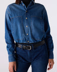 A model wearing a cropped, long-sleeve blue denim shirt with buttoned chest pockets, tucked into dark blue jeans with a black belt, against a plain light gray background.