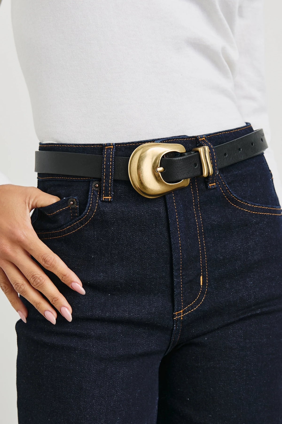 Dark blue jeans with a gold belt buckle worn by a person on a light gray background