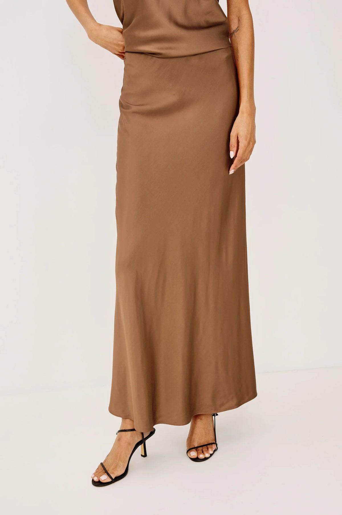 A woman wearing a long, flowing taupe-brown satin maxi skirt with a matching top and black strappy high-heeled sandals, photographed against a clean off-white studio background.