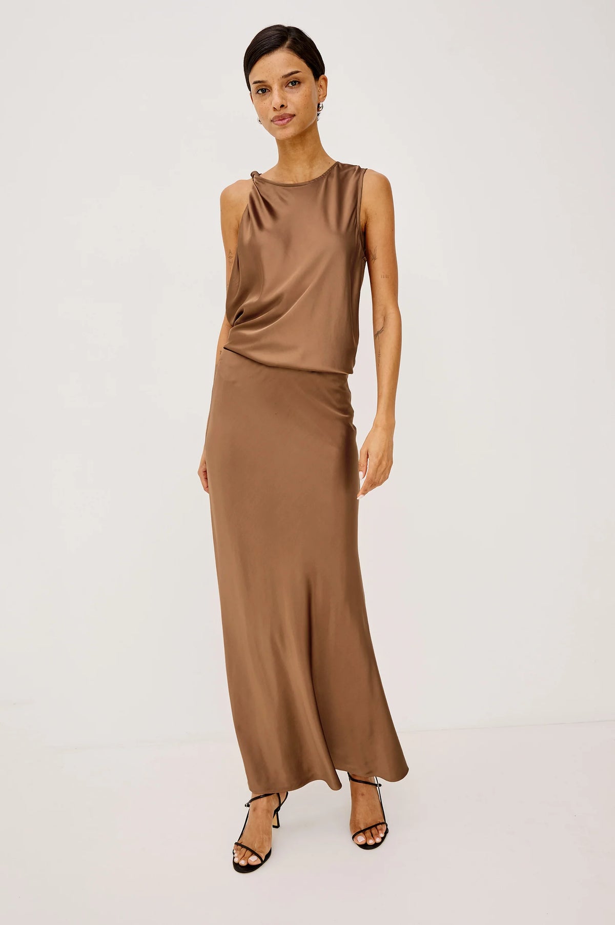 A woman modeling a sleeveless, high-neck, two-piece satin dress in a warm taupe-brown color, paired with black strappy heels, against a plain white studio background.