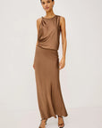 A woman modeling a sleeveless, high-neck, two-piece satin dress in a warm taupe-brown color, paired with black strappy heels, against a plain white studio background.
