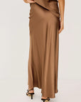 A woman wearing a long, flowing taupe-brown satin maxi skirt with a matching top and black strappy high-heeled sandals, photographed against a clean off-white studio background viewed from the back 
