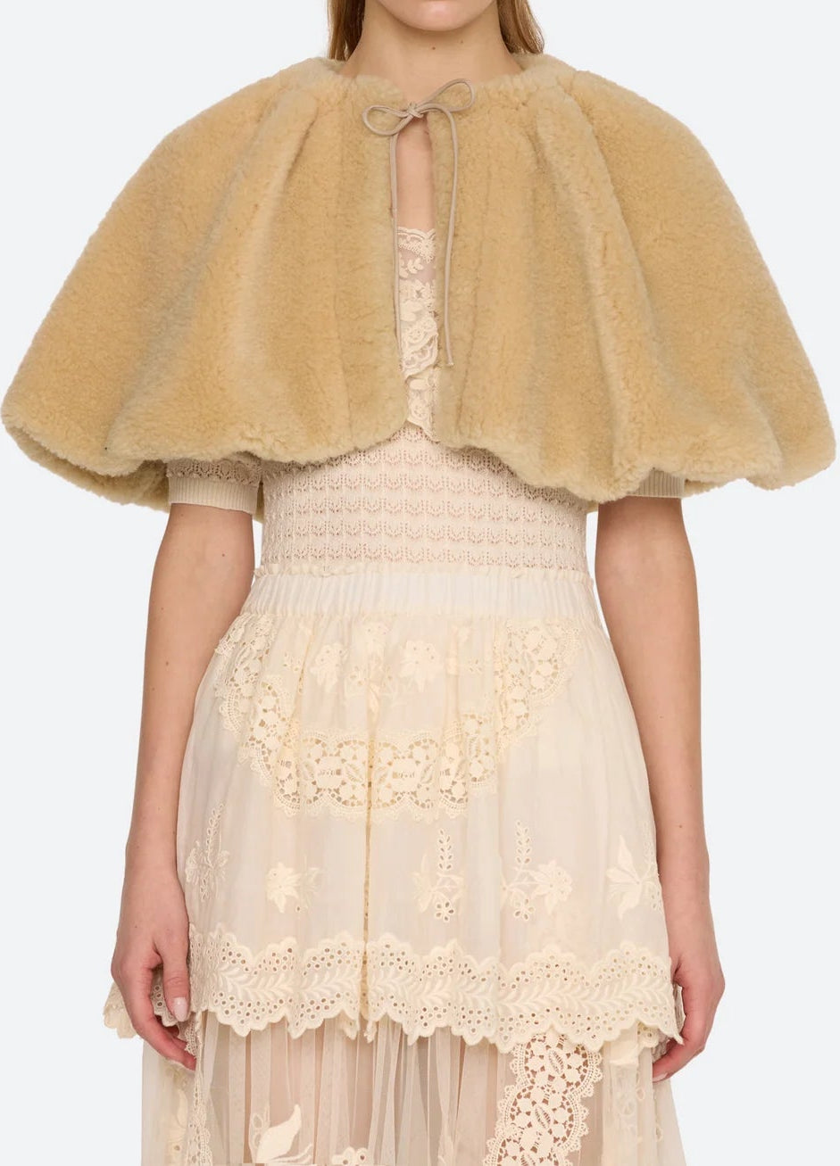 A model wearing a tan fuzzy faux-fur cape with a tie-neck detail, layered over a cream lace dress, against a light gray background.