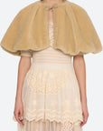 A model wearing a tan fuzzy faux-fur cape with a tie-neck detail, layered over a cream lace dress, against a light gray background.