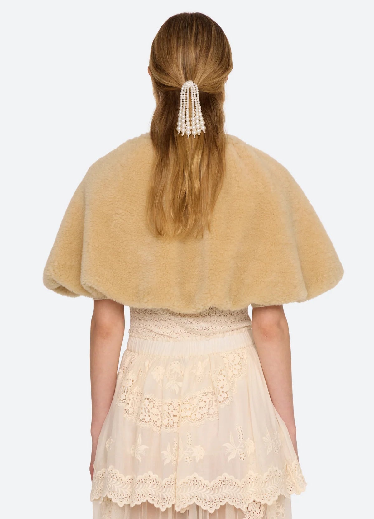 A model wearing a tan fuzzy faux-fur cape with a tie-neck detail, layered over a cream lace dress, against a light gray background, back view