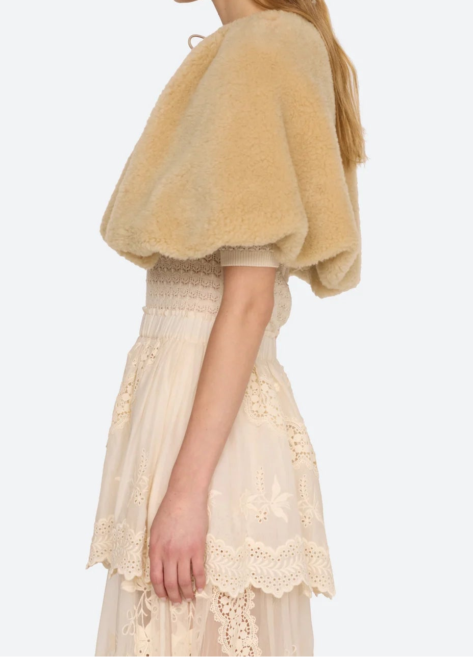 A woman shown in side profile wearing a fuzzy short cape-style jacket in a warm beige/tan color over a cream lace dress, against a plain light gray studio background.