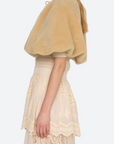 A woman shown in side profile wearing a fuzzy short cape-style jacket in a warm beige/tan color over a cream lace dress, against a plain light gray studio background.