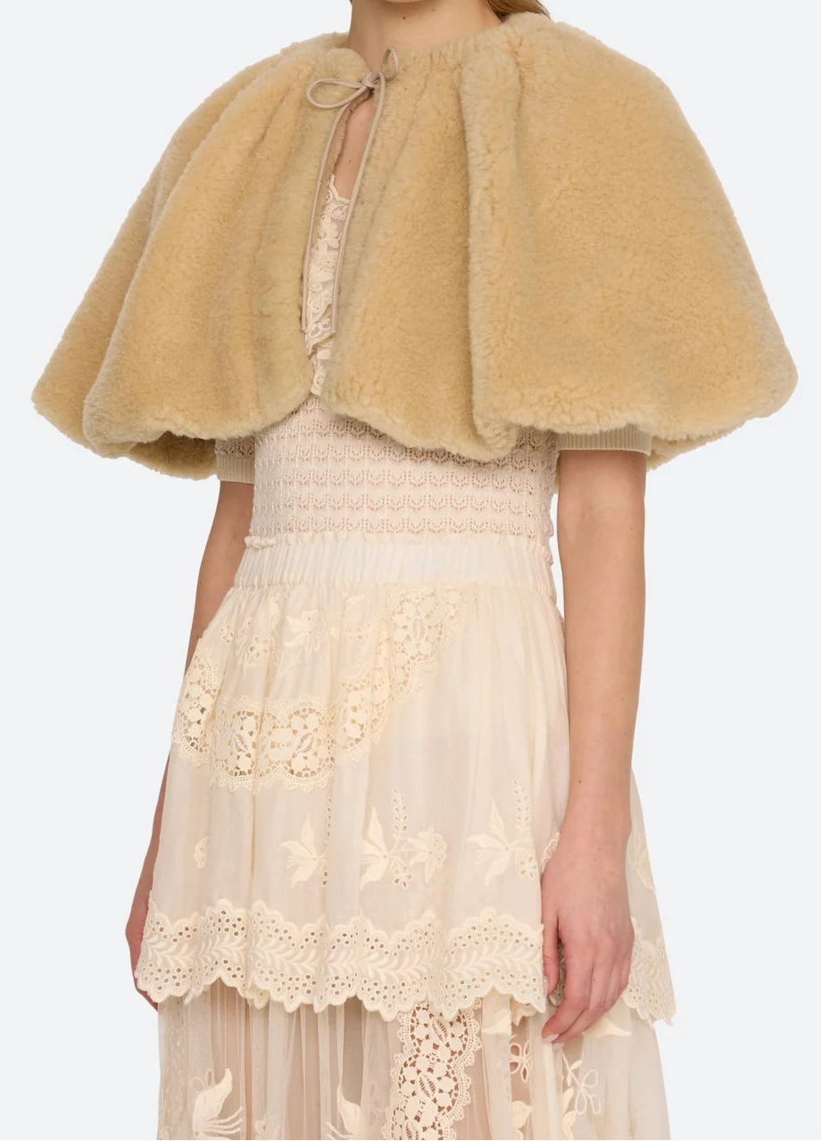 A model wearing a beige fuzzy cropped cape over a cream lace dress, photographed against a light gray studio background.