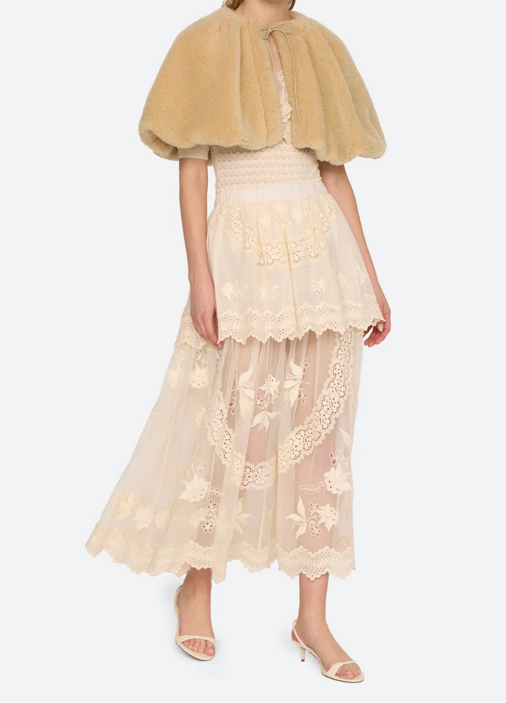 A model wearing a beige faux-fur capelet over a cream lace tiered midi skirt and nude high-heeled sandals, set against a plain light gray studio background.