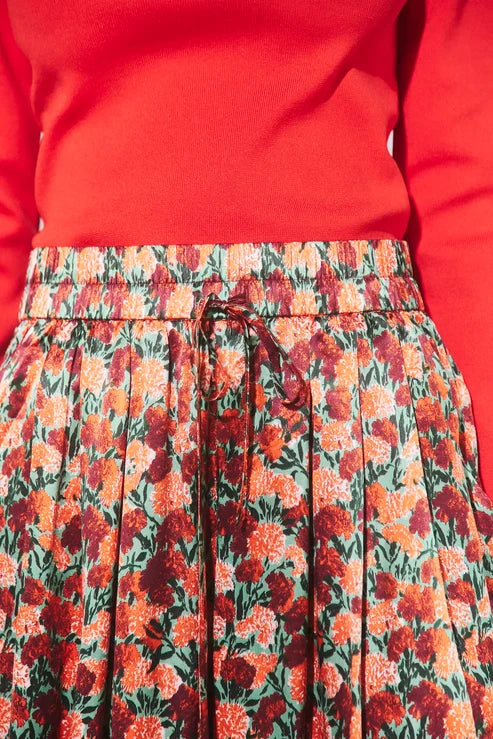 Close-up of a pleated skirt with a red, orange, and green floral print and a drawstring waist, paired with a bright red top.
